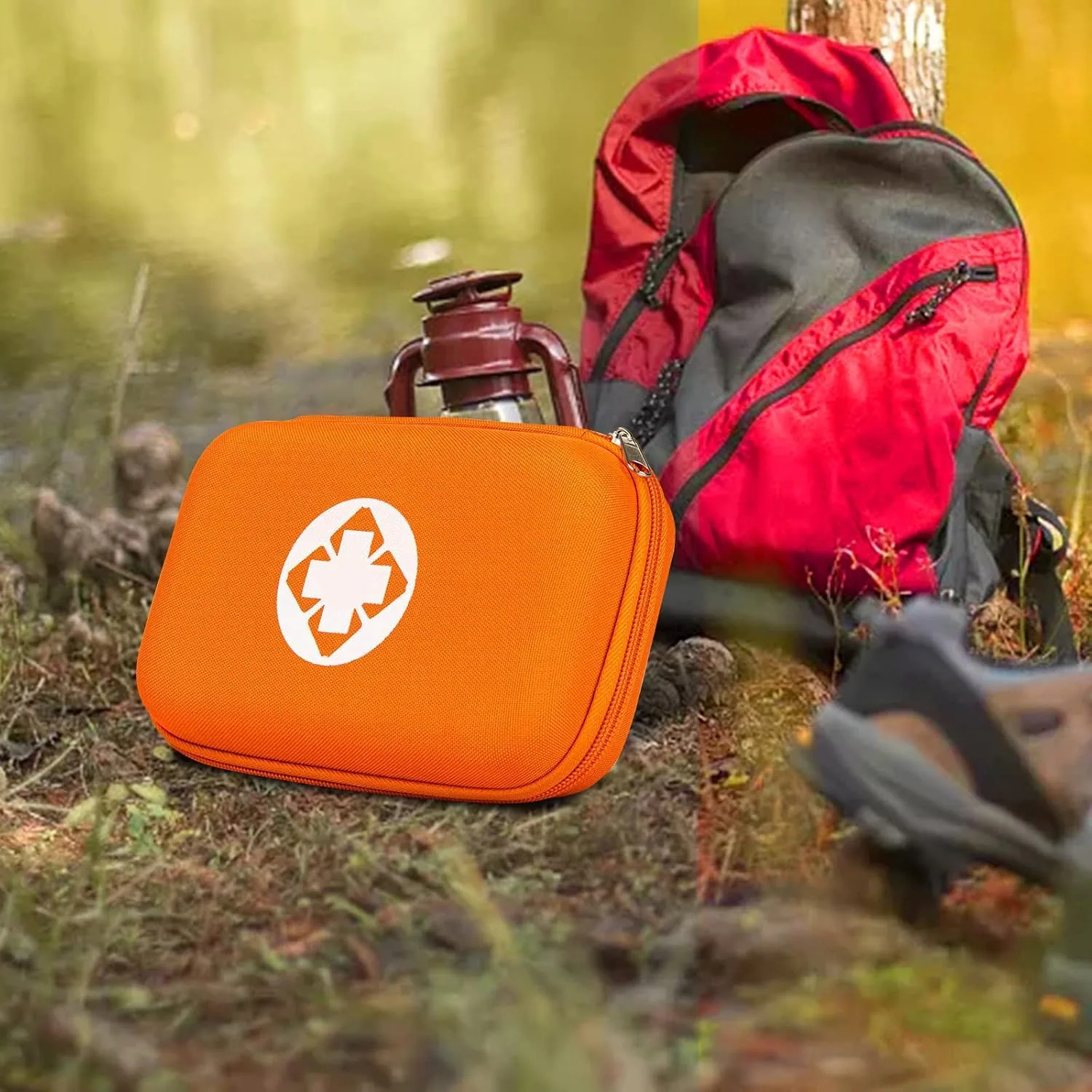 An orange AMORNING first aid kit by a red backpack and a lantern on a forest floor